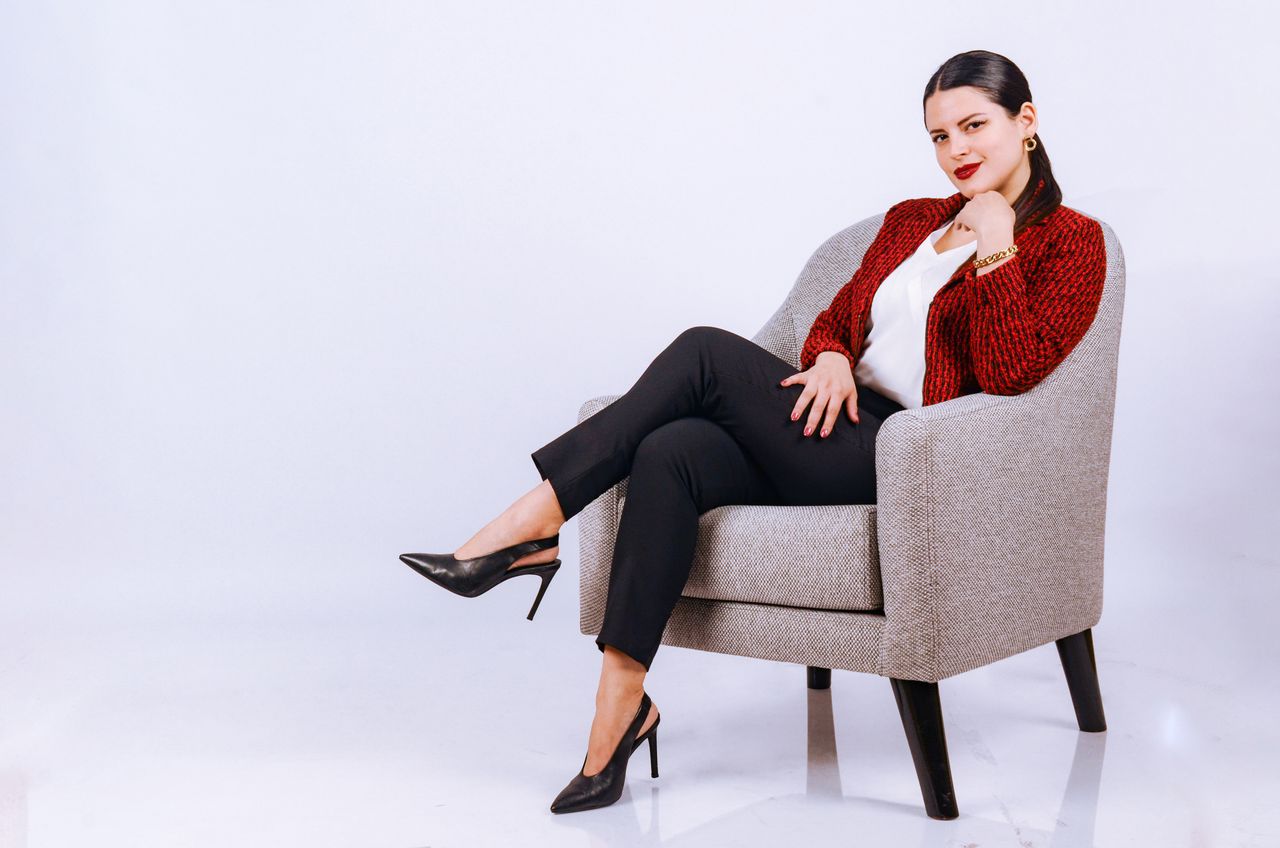 Fashionable woman in a red jacket and heels sitting on a stylish armchair against a white background.